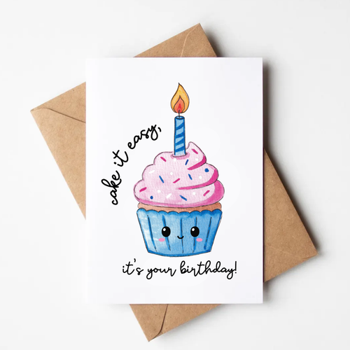 A white greeting card featuring a cute watercolor-style cupcake with pink frosting, a blue wrapper, a single lit candle, and the text 'cake it easy, it's your birthday!' in black script. The card is photographed on a light neutral background with a brown kraft envelope behind it.