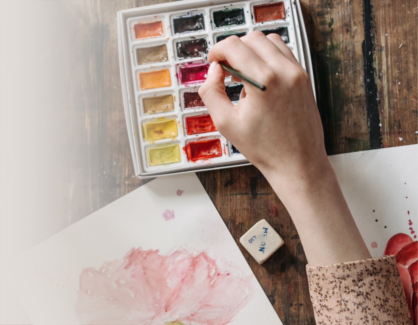 A hand using a paintbrush to mix colors in a 24-color watercolor paint palette, with painted paper sheets on a rustic wooden table.