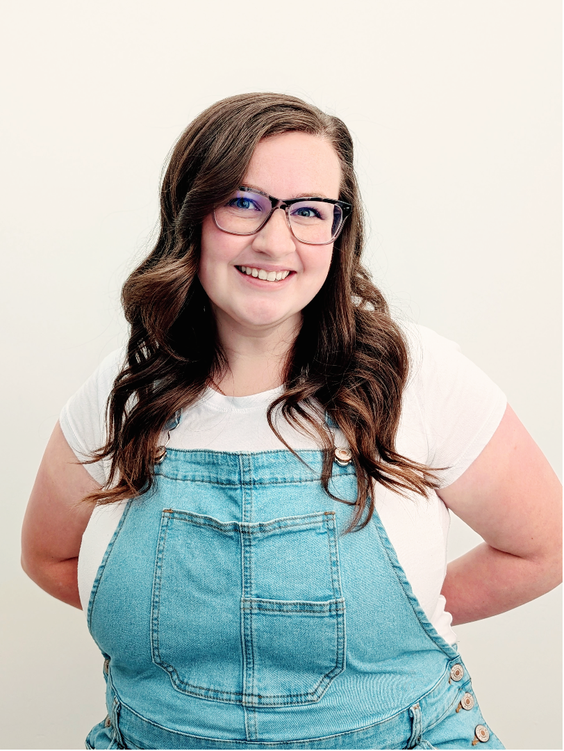 A smiling woman with long brown hair and glasses, wearing a white t-shirt and light blue denim overalls, posing against a plain off-white background.